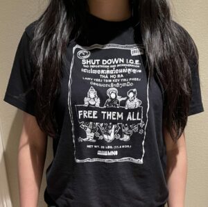 A black t-shirt with a rice bag design that says, “Shut Down ICE. End deportation and incarceration. Free Them All.” On the rice bag design, there are three ladies dressed in various traditional Southeast Asian clothing, and “Free Them All” is written in various Southeast Asian languages.