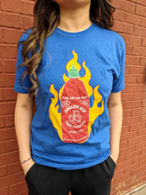 A blue t-shirt with a red sriracha bottle on fire that says, “Turn up the heat. Abolish ICE. Free Them All.”