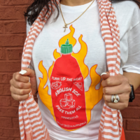 A white t-shirt with a red sriracha bottle on fire that says, “Turn up the heat. Abolish ICE. Free Them All.”