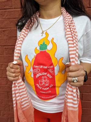 A white t-shirt with a red sriracha bottle on fire that says, “Turn up the heat. Abolish ICE. Free Them All.”